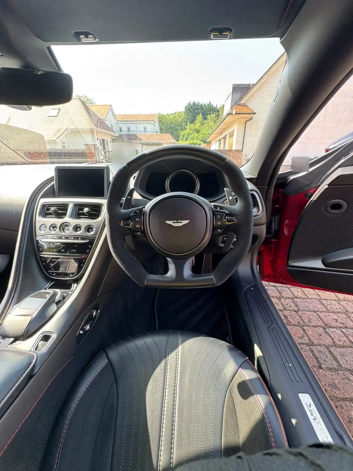 Aston Martin DB11 cabin seen through the open driver door, showing the steering wheel, dashboard and black leather seat with red stitching
