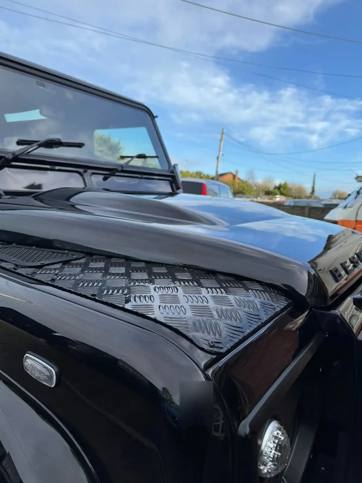 Close up of a black Land Rover Defender bonnet and wing, showing sky reflections in the paint and the chequer plate on the wing top