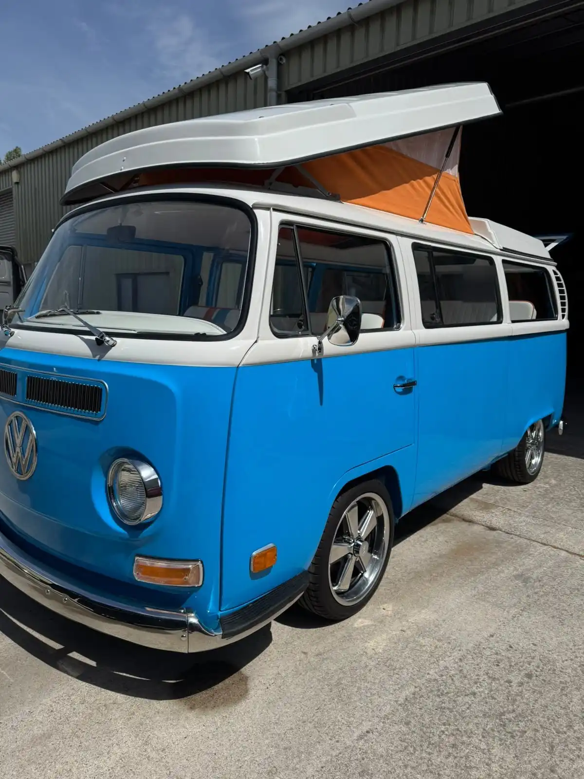 Blue and white VW T2 camper from the front three quarter with the pop top raised, showing glossy paintwork and bright alloy wheels