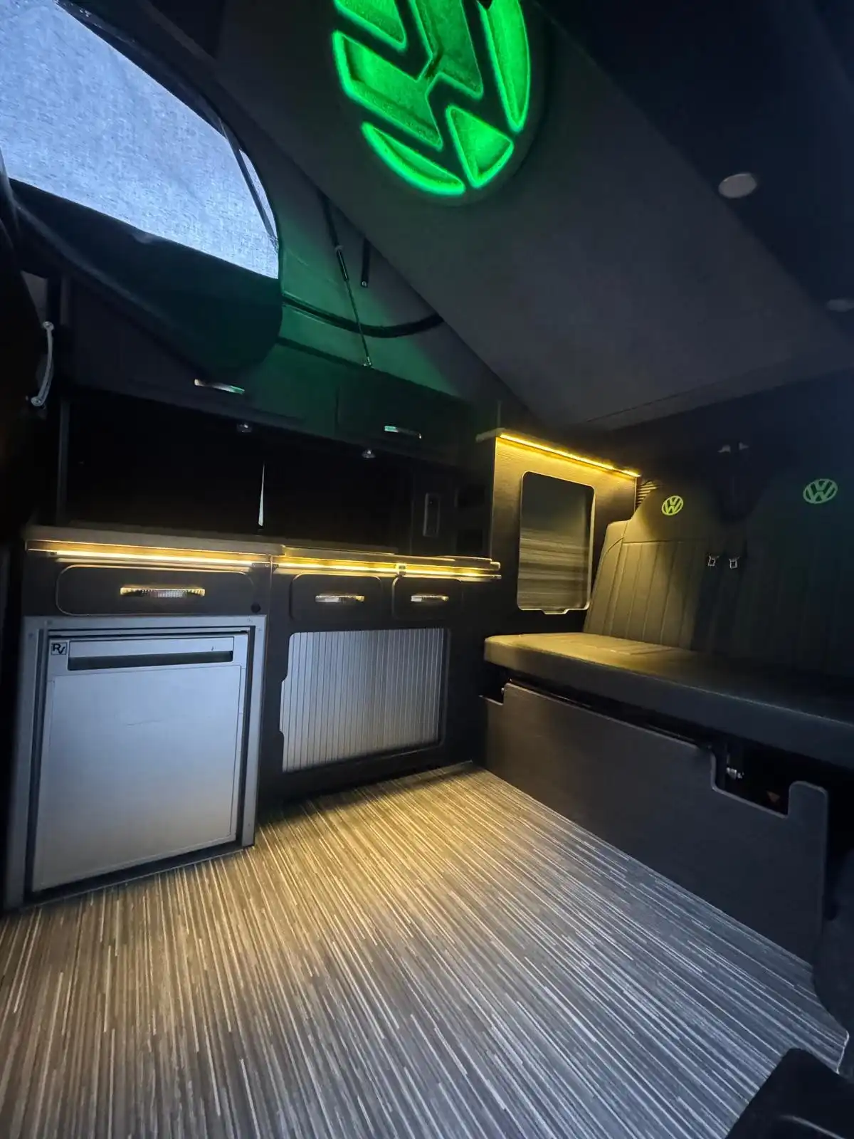 Inside a VW Transporter camper conversion, showing the units, fridge and bench under warm strip lighting
